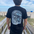 Person wearing a black t-shirt with a graphic design on the back, standing on a wooden boardwalk.