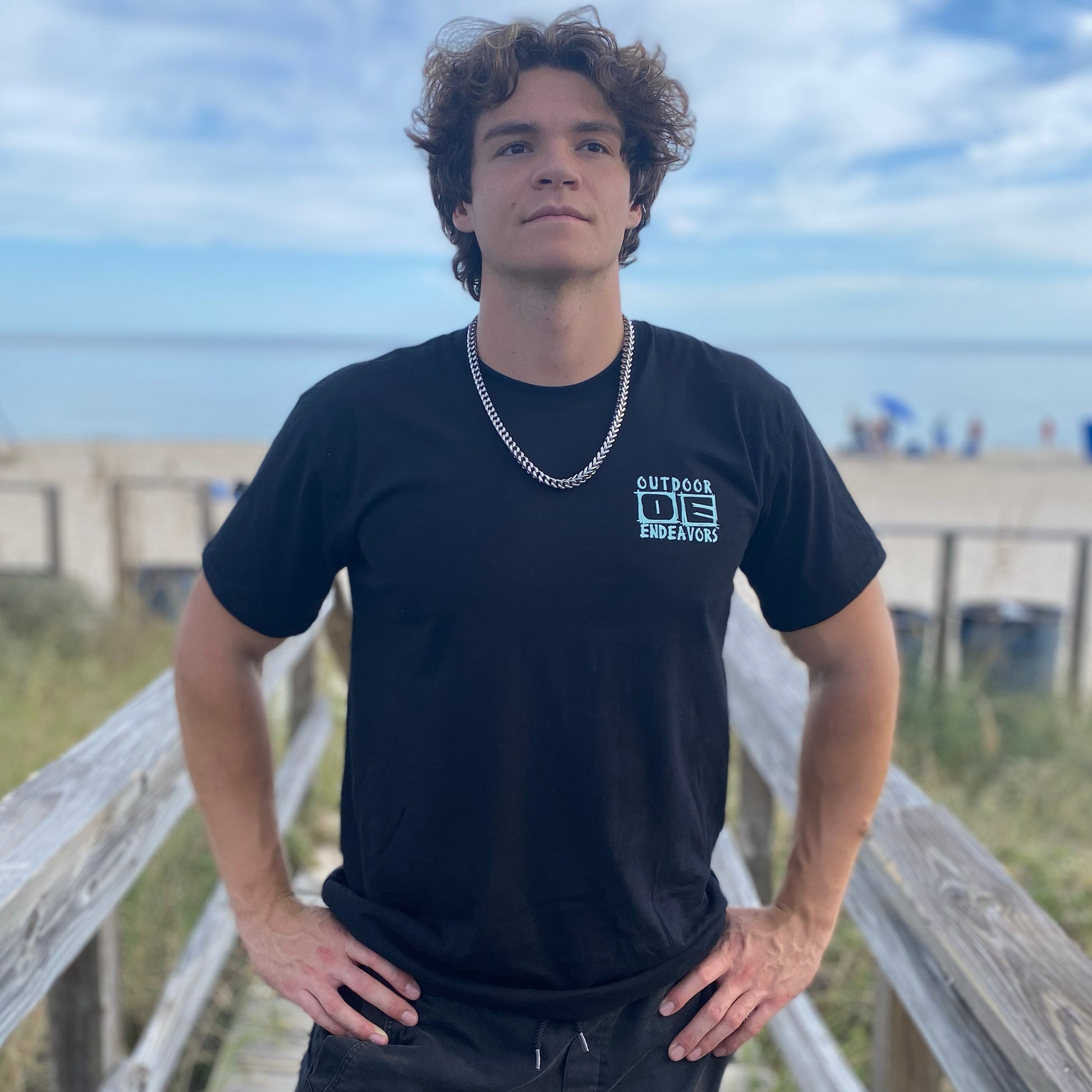 Person wearing a black t-shirt with text on a wooden boardwalk with grass and sky in the background