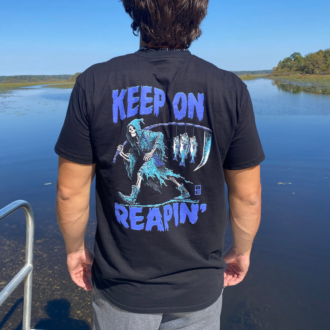 Black t-shirt with blue graphic of a skeleton holding a scythe and text 'KEEP ON REAPIN'' on the back, brand logo on the front.