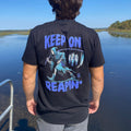 Person wearing a black t-shirt with a graphic design and text by a body of water.