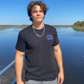 Person wearing a black t-shirt with a logo standing by a body of water.