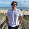 Person wearing a white t-shirt with text standing on a wooden boardwalk by the beach.